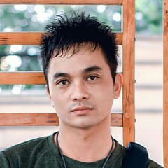 Young adult man holds a book and bag in a casual outdoor setting, appearing pensive.